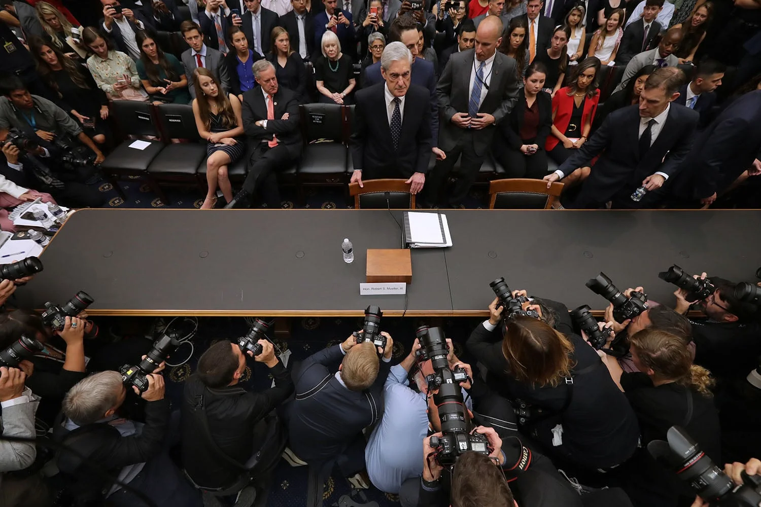 News photographers point their cameras at Robert Mueller as he stands in front of a chair before testifying. A crowd of people are seen behind him.