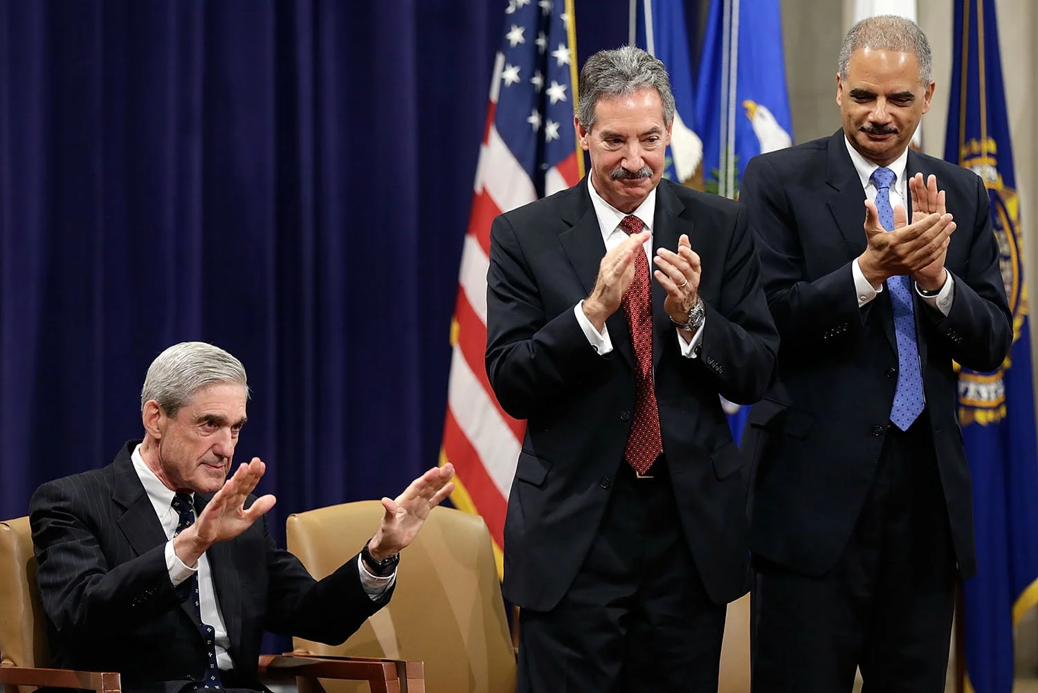 Robert Mueller, seated, holds both hands out in a gesture of humility as two other man stand next to him while clapping. Behind them are flags of the United States and other agencies.
