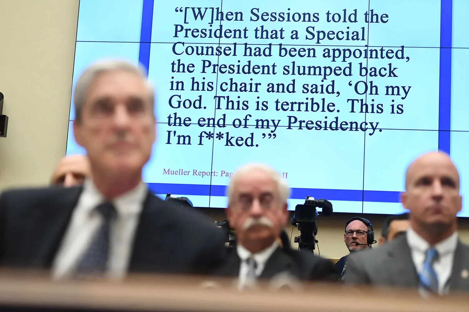 Robert Mueller is seen out of focus in the foreground along with other men. A large screen behind them reads: "[W]hen Sessions told the President that a Special Counsel had been appointed, the President slumped back in his chair and said, 'Oh my God. This is terrible. This is the end of my Presidency. I'm f**ked.'"