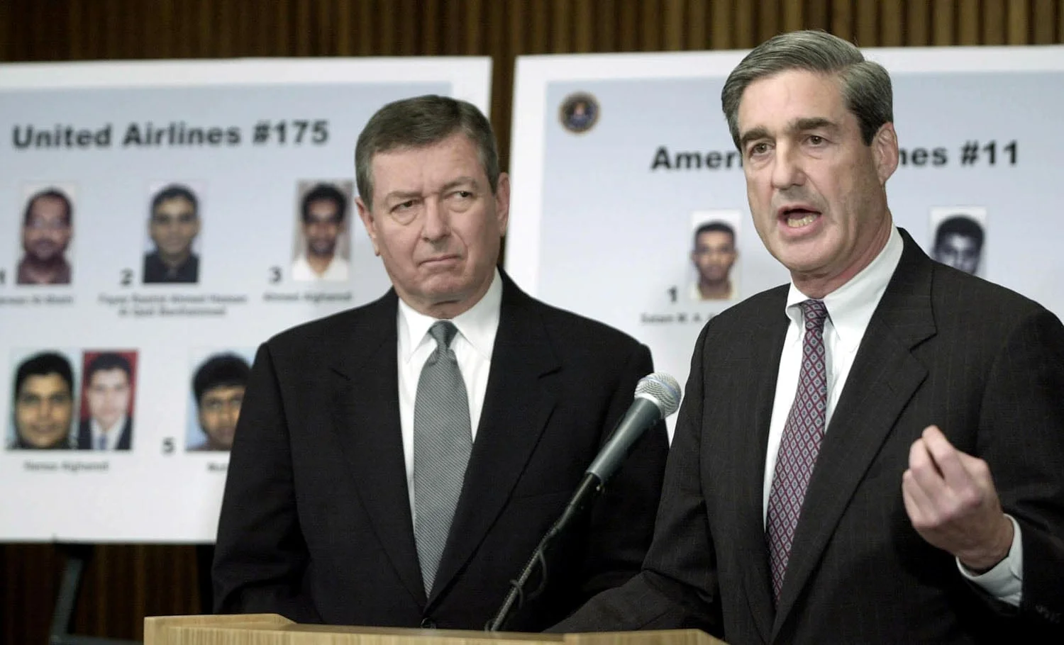 Robert Mueller speaks behind a microphone as John Ashcroft watches. Behind them are poster boards with headshots of men and labels: United Airlines #175 and American Airlines #11.