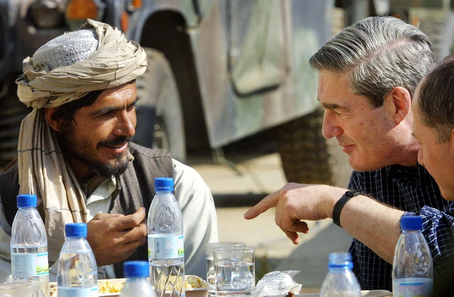 A man on the left points with one hand into frame in front of Robert Mueller, who smiles slightly as he leans toward Haji Gulalai, who is also smiling and wearing a turban. Water bottles and glasses sit in front of them on a table. A military vehicle is out of focus behind them.