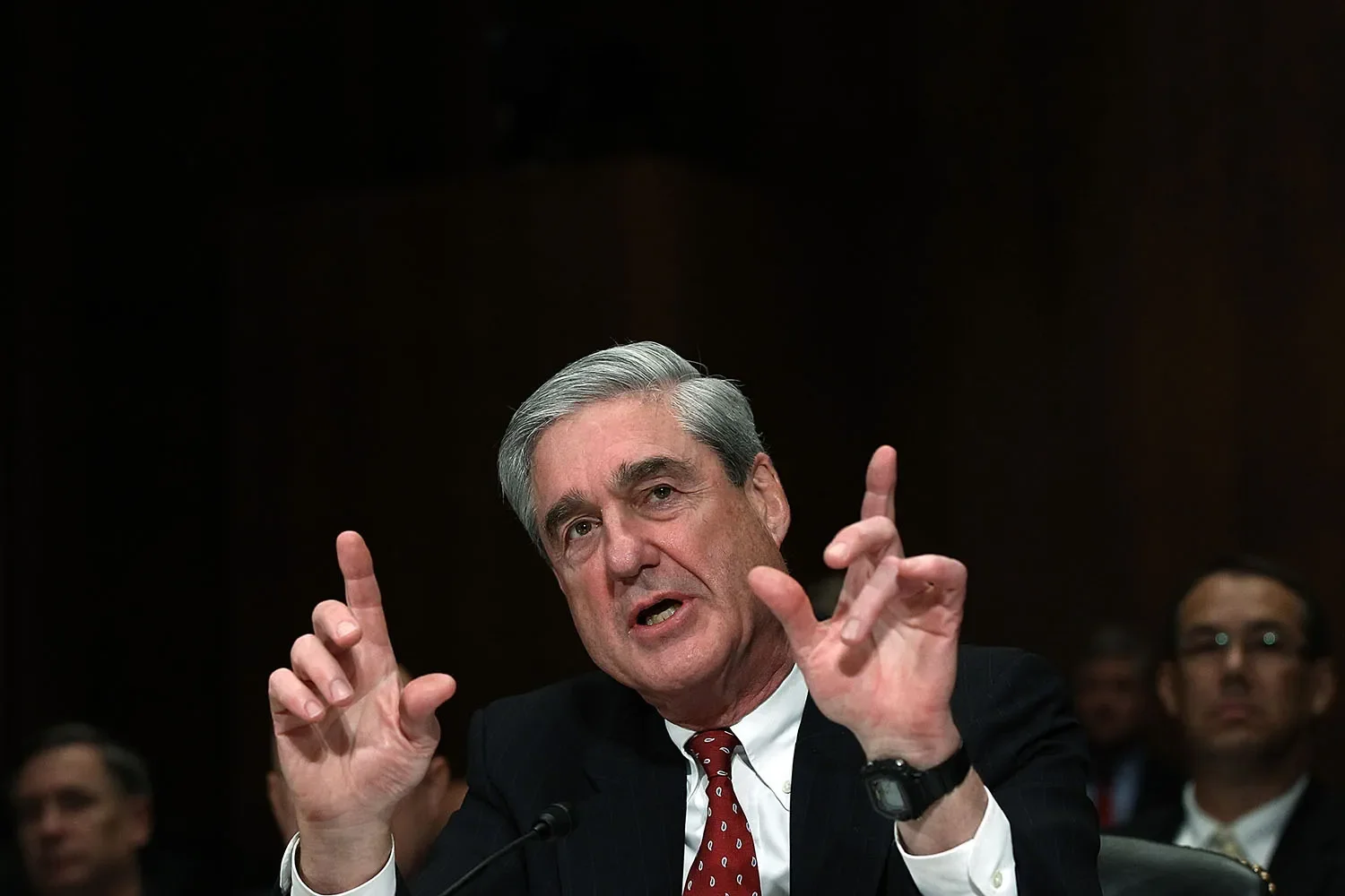 Robert Mueller gestures with both hands as he talks in front of a microphone.