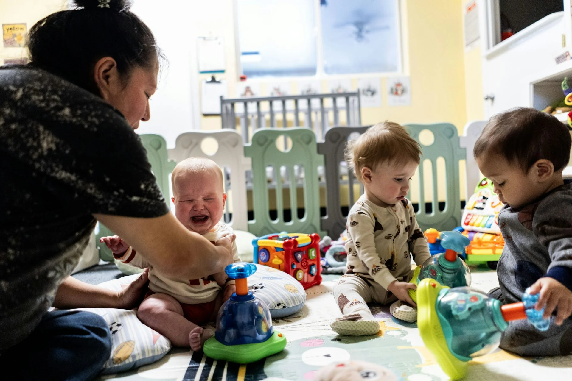 A woman leans over three babies and toddlers who are sitting on the floor among plastic toys. One of them is crying.