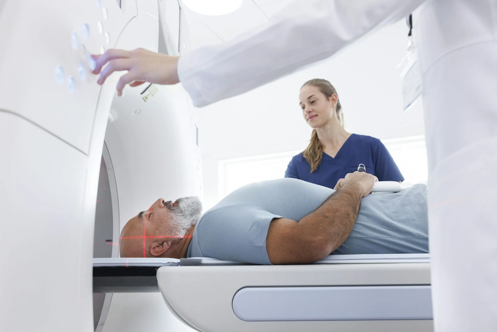 A man lies on his back, entering a CT scanner, with two health professionals overseeing him.