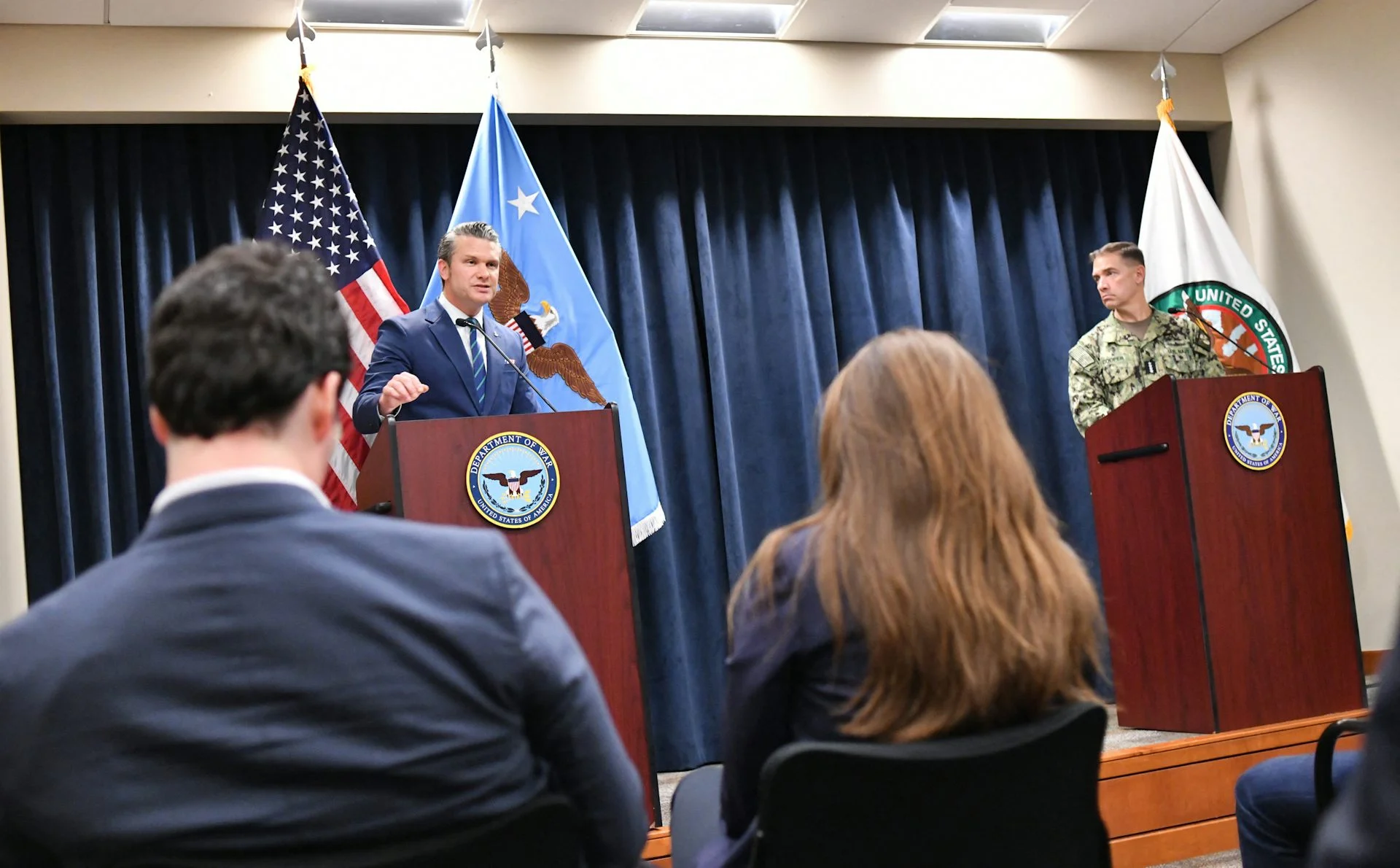 Two men, including one in a military uniform, at lecterns, speaking.