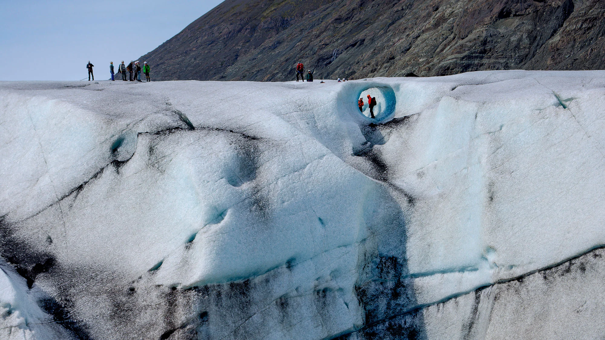 A group of tourists stands on top of a glacier, with two framed underneath a rounded arch in the side.