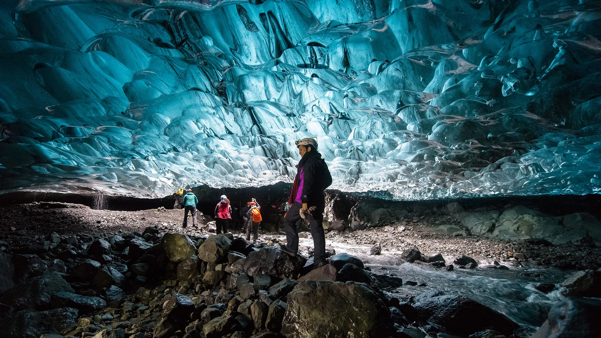 a group of tourists in coats and helmets stand inside a cave, beneath an impressive shiny blue and silver wall of ice