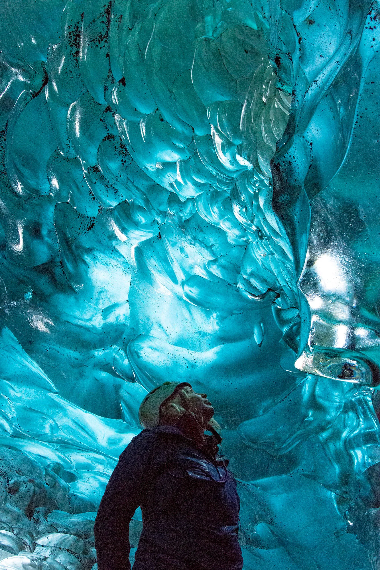 A woman stands beneath a bubbly cave wall of crystal blue ice, looking up