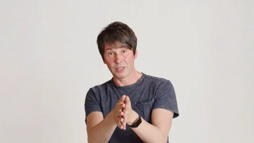 A man with short dark hair, wearing a dark t-shirt and smartwatch, gestures with his hands while standing in front of a plain white background.