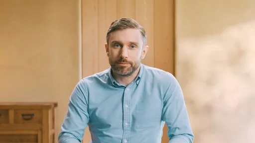 A man with short hair and a beard, wearing a light blue button-up shirt, sits indoors in front of a wooden wall.