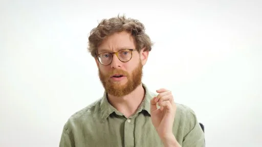 A man with curly hair, a beard, and glasses, wearing a green button-up shirt, gestures with his hand against a plain white background.