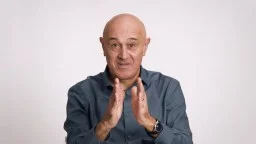 Bald man in a blue shirt gestures with both hands in front of him, palms facing each other, against a plain white background.