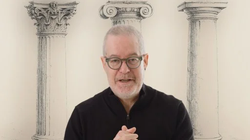 A man with glasses and a gray beard stands in front of illustrations of three classical columns, each with a different capital style.