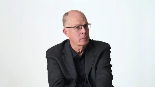 Bald man wearing glasses and a dark suit jacket sits against a plain white background, looking slightly to the side with a neutral expression.