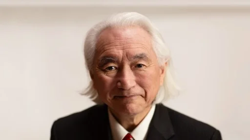 An older man with long white hair wearing a dark pinstriped suit, white shirt, and red tie, looking directly at the camera against a plain light background.