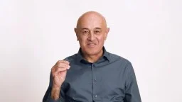 Bald man in a dark button-up shirt gestures with his right hand while looking at the camera, against a plain white background.