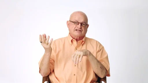 Bald man wearing glasses and a peach shirt sits on a chair against a white background, gesturing with his left hand while talking.