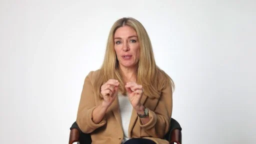 A woman with long blonde hair sits on a chair against a plain white background, wearing a tan jacket and gesturing with both hands while speaking.