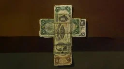 A cross made from various denominations of old U.S. paper currency is fastened together with brass tacks, set against a brown background.
