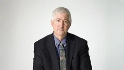 An older man with gray hair wearing a dark suit, blue shirt, and patterned tie, sitting against a plain light background.