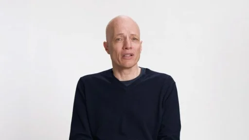 A bald man wearing a dark sweater sits against a plain white background, looking slightly to the side with a neutral expression.