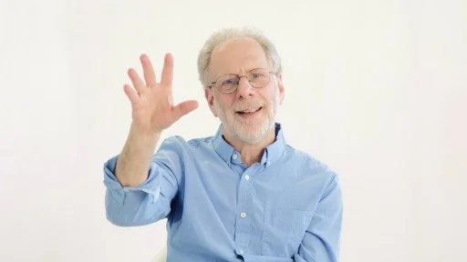 An older man with glasses and a light blue shirt sits and smiles while raising his hand with fingers spread, in front of a plain white background.