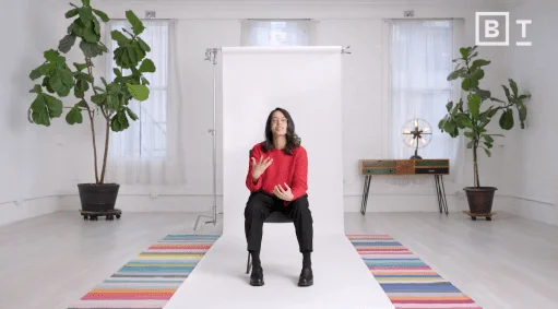A person in a red sweater sits on a stool in front of a white backdrop in a bright room with plants, a colorful rug, and a table with a lamp.