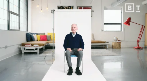 An older man sits on a chair in front of a white backdrop in a modern, brightly lit room with colorful pillows and minimalist decor.