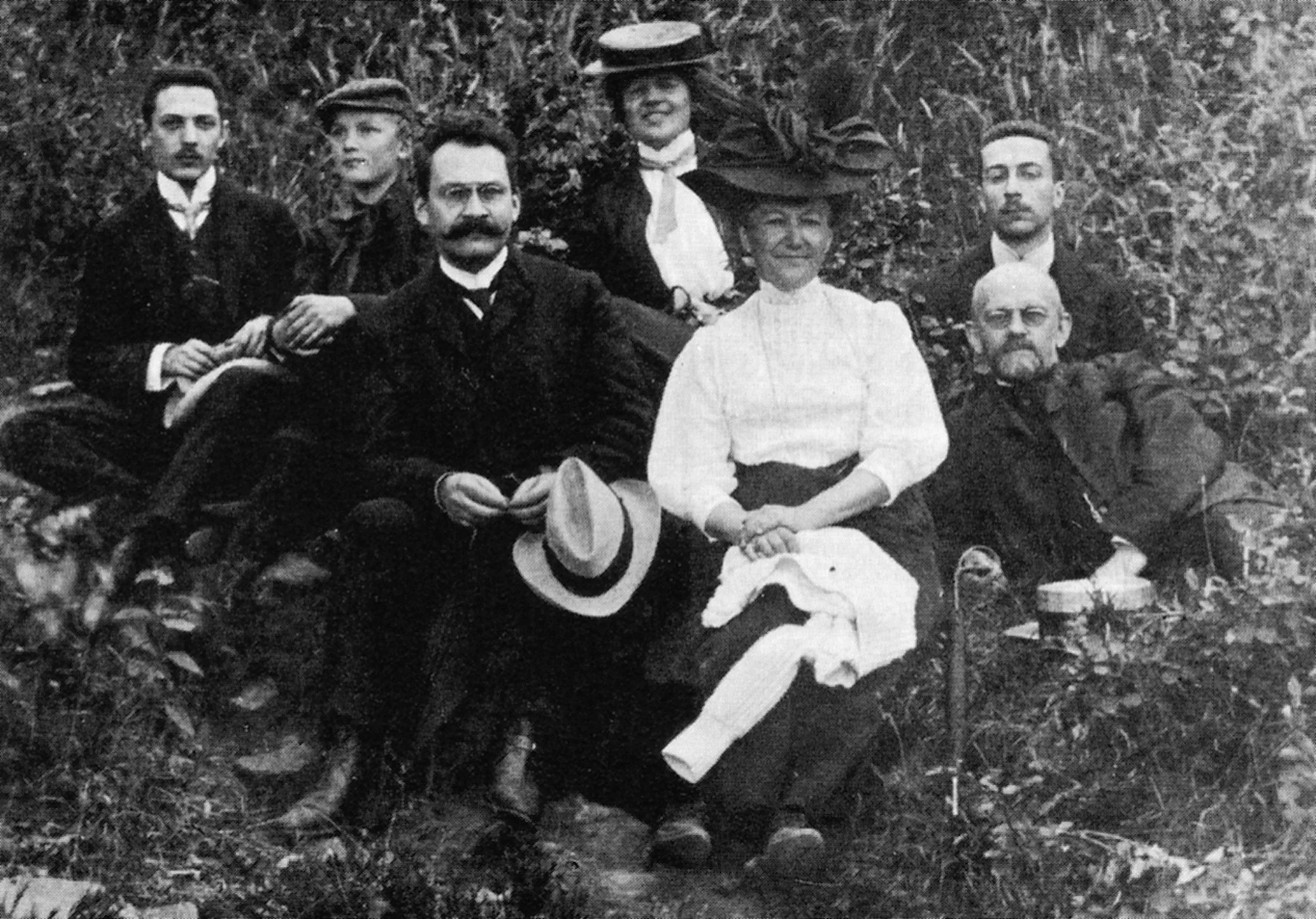 A group of seven people, four men and three women, sit outdoors on grass in formal late 19th or early 20th century clothing, posing for a photograph that captures the science value of documenting history before it is destroyed by time.