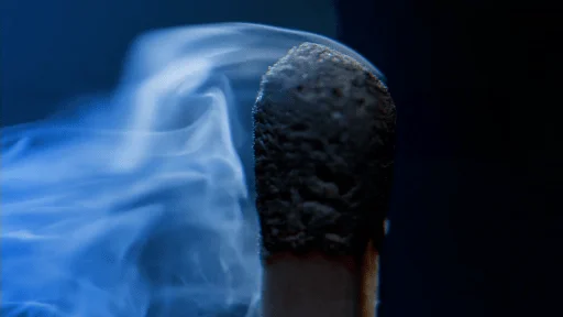 Close-up of a burnt matchstick with smoke swirling around its charred tip against a dark background.
