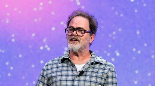 A bearded man wearing glasses and a plaid shirt speaks with a microphone clipped to his collar in front of a purple background with white dots.